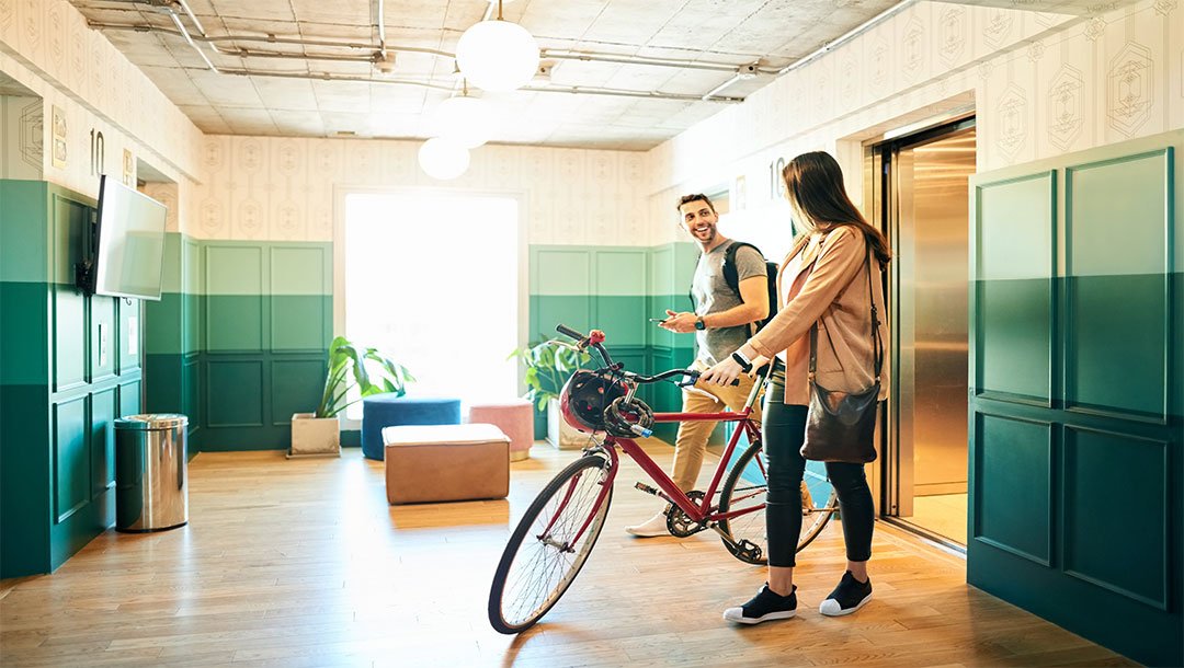 Two people with a bicycle talking near an open elevator in a modern lobby