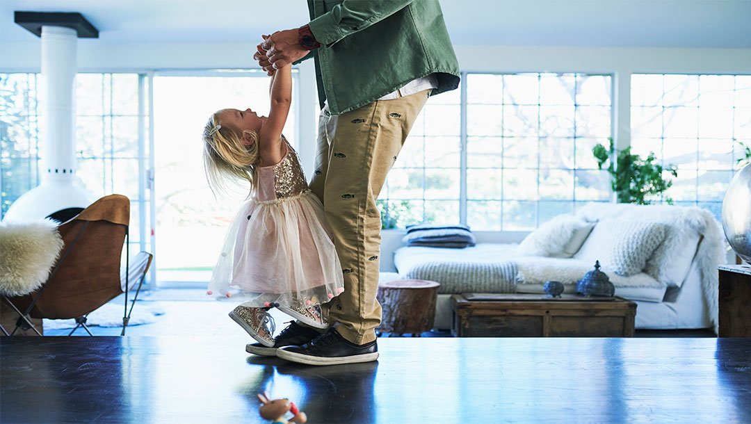 Small child holding an adult’s hands while standing on their feet in a bright living room
