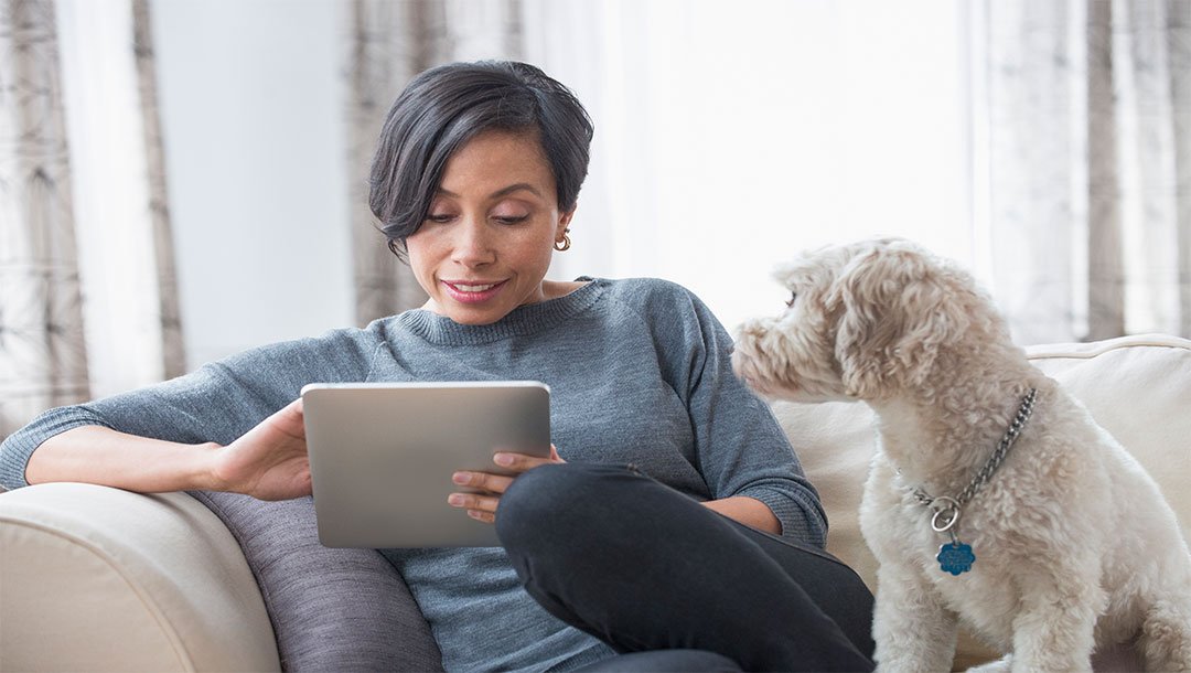 Woman sitting on sofa reading from a tablet while dog looks on